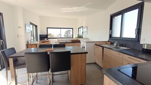 Een moderne keuken met een centraal eiland, zwarte barkrukken, houten kastjes en een groot raam dat voor natuurlijk licht zorgt.
