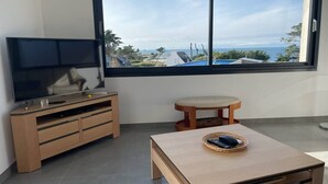Een kamer met een televisie op een houten kastje, een klein houten tafeltje met een afstandsbediening, en een groot raam met uitzicht op zee en gebouwen.