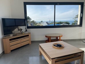 Una stanza con una TV su un mobile in legno, un piccolo tavolino con un telecomando e un'ampia finestra vista mare e palazzi.