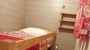 Ein Stockbett mit einer roten und weißen Bettdecke, einem kleinen Holzregal und einer Metalttür.