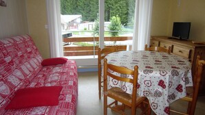 Ein Zimmer mit einem roten, gemusterten Sofa, einem Esstisch mit weißer Tischdecke, Holzstühlen und einem Fernseher auf einer Holzkommode.