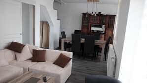 Een moderne woonkamer met een bank, een salontafel en een zwarte fauteuil. Achtergrond toont een eethoek met een tafel en stoelen.
