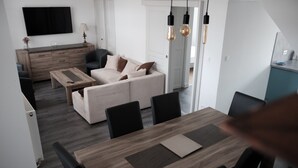 Een moderne woonkamer met een bank, salontafel en eettafel. Er is een televisie aan de muur bevestigd, een lamp en een kast.