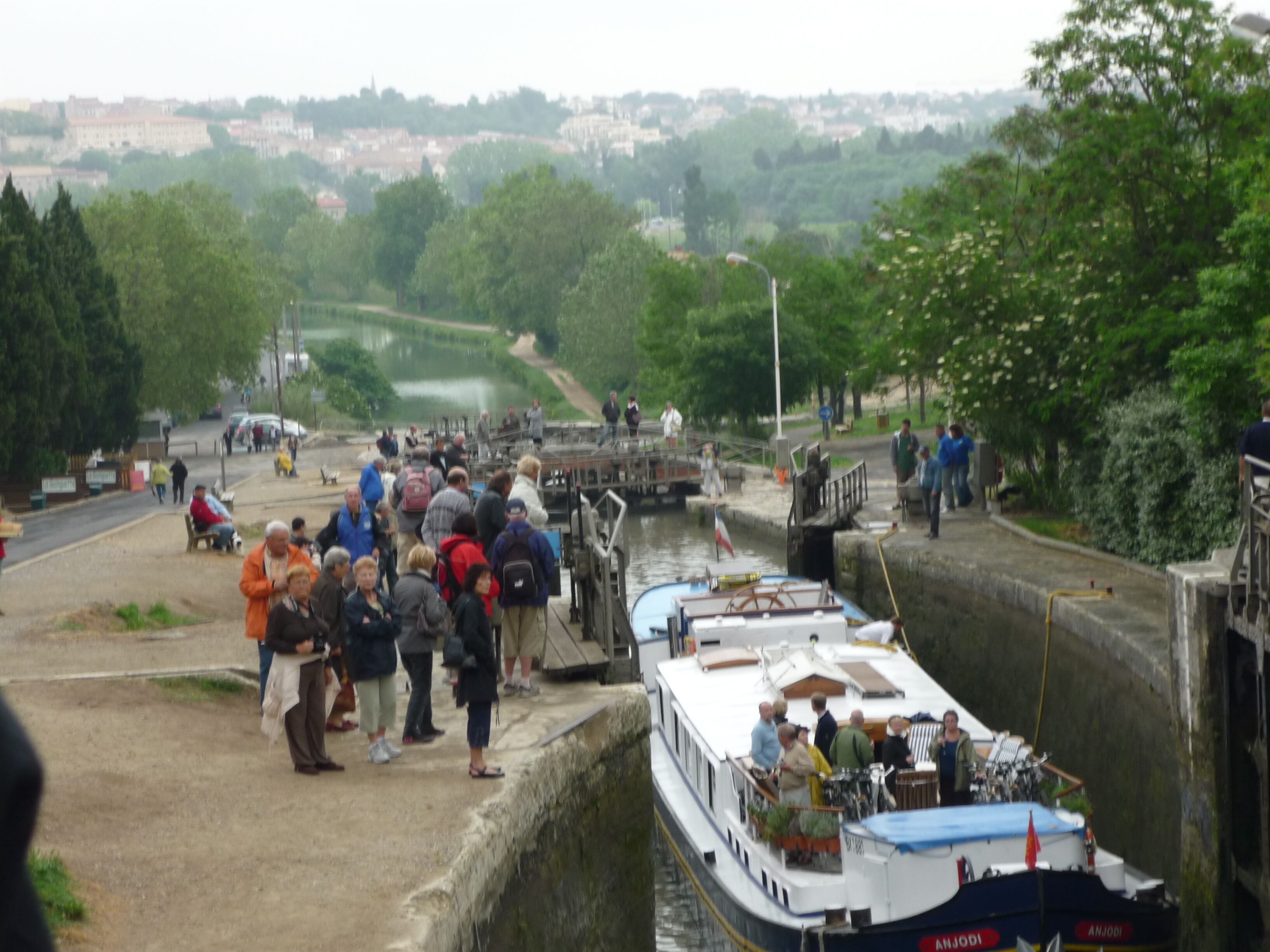 Un groupe de personnes s’est rassemblé près d’un canal où étaient amarrés des bateaux. On pouvait apercevoir un pont en arrière-plan et, au loin, le paysage urbain.