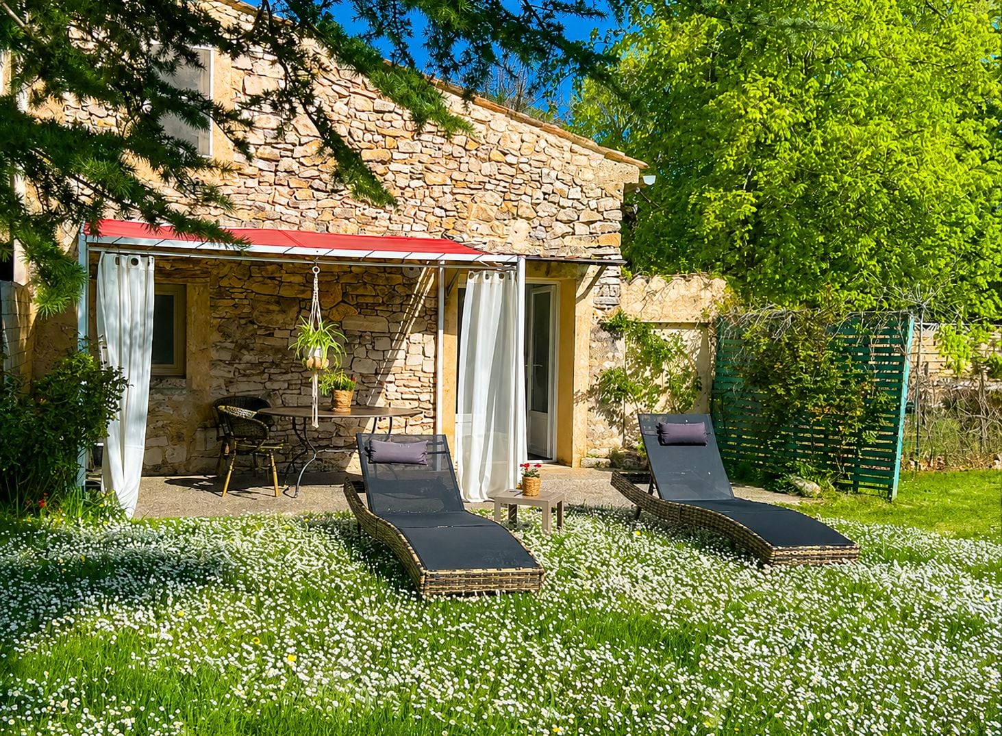 A stone building with a covered patio, two lounge chairs, a table with chairs, and a garden with white flowers.