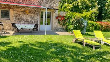 Une maison en pierre avec un patio couvert, une table et des chaises, ainsi que deux fauteuils de jardin jaunes.