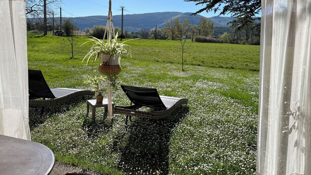 Une terrasse couverte avec une table et des chaises, une plante suspendue, et une vue sur un champ verdoyant avec des arbres et des collines en arrière-plan.