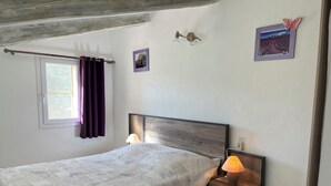 Une chambre à coucher avec un lit, une table de chevet avec une lampe, une fenêtre avec des rideaux et des tableaux accrochés au mur.