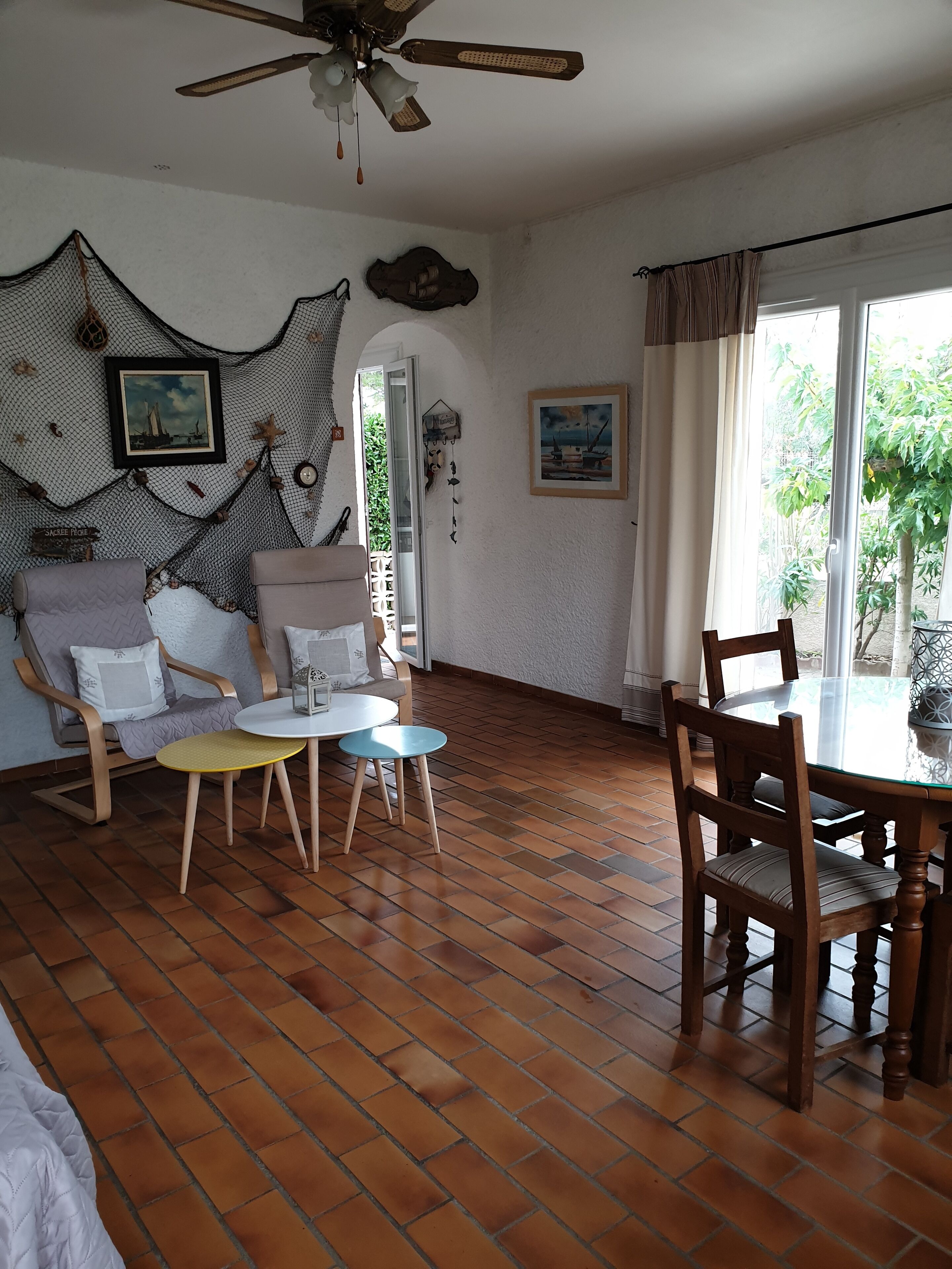 A living room with a wooden floor, a ceiling fan, a dining table with chairs, and a large window with curtains.