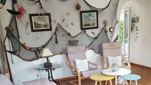 A cozy living room with a cushioned sofa, a small round table, and two chairs. A decorative net hangs on the wall, and there are framed pictures above the sofa.