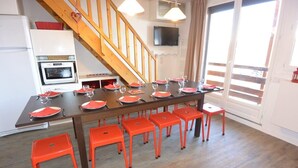 A dining area with a long table set for six, surrounded by six orange chairs, a wooden staircase, a television mounted on the wall, and a sliding glass door leading to a balcony. - Risoul 4 bedroom duplex sleeps 11, at the foot of the slopes, summer winter (Risoul)