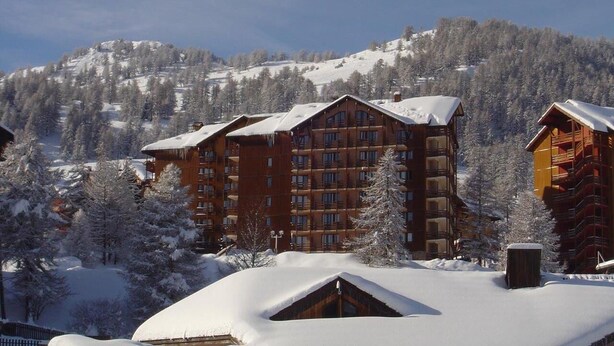 A lodge with multiple wooden buildings, surrounded by snow-covered trees and mountains in the background. - Risoul 4 bedroom duplex sleeps 11, at the foot of the slopes, summer winter (Risoul)