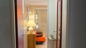 A hallway leading to a bedroom with a bed, bedside table, lamp, and a small shelf with items.