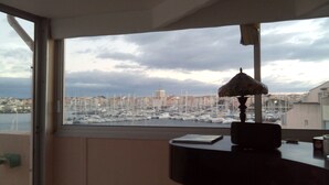 Een kamer met een groot raam met uitzicht op een jachthaven vol boten, met een stadsgezicht op de achtergrond en een tafel met een lamp.