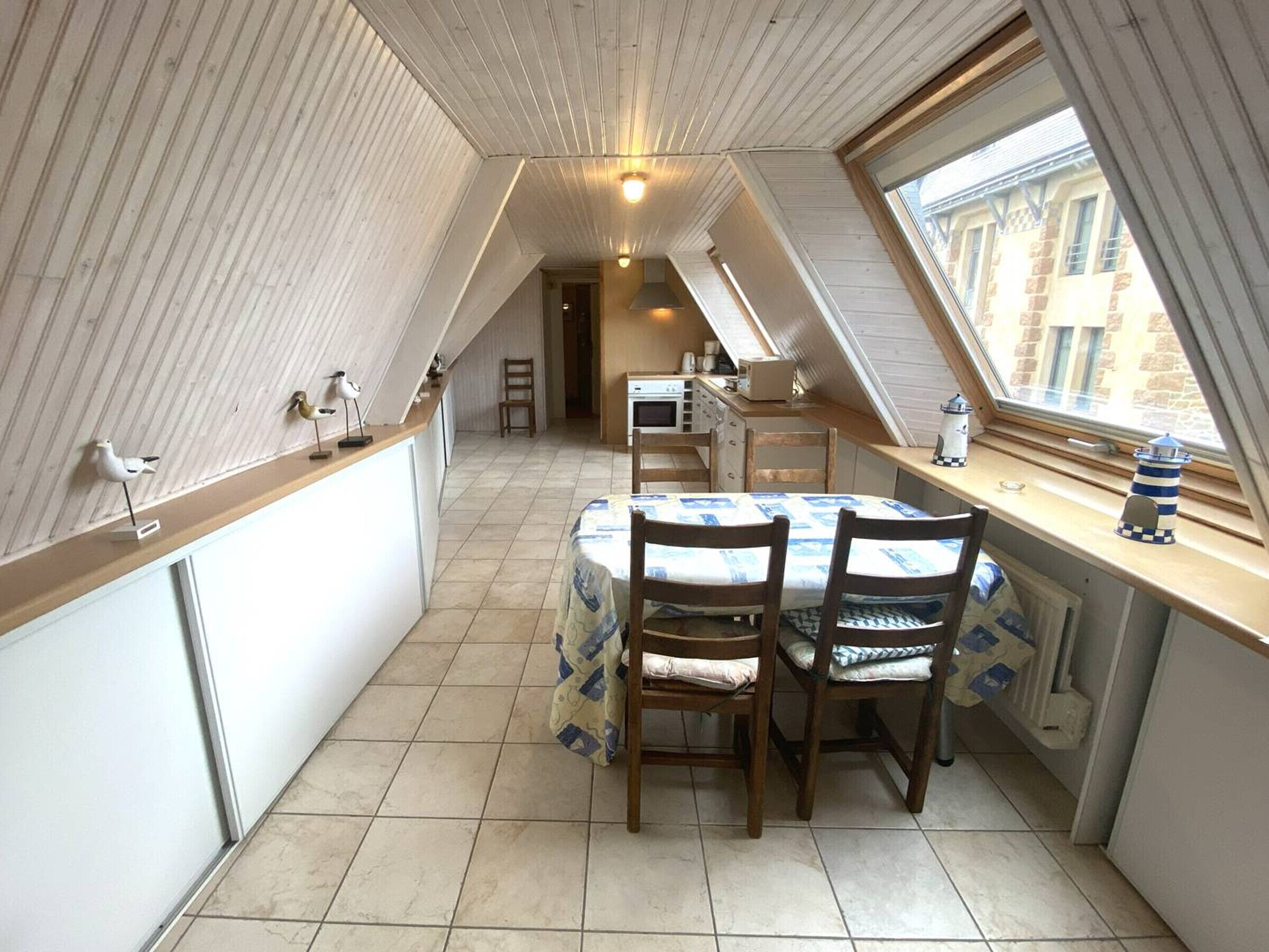 A compact kitchen with a dining area, featuring a small table and chairs, a window with a view of buildings, and a ceiling with sloped wooden panels.
