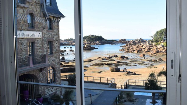 Une vue depuis une fenêtre d’hôtel donnant sur une plage rocheuse, avec une pancarte indiquant « Hôtel Beau Site ».