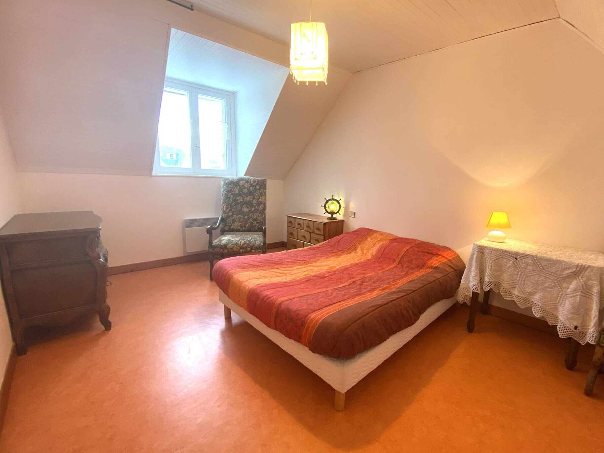 Een slaapkamer met een bed, een stoel, een dressoir, een lamp en een raam.