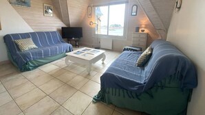 Une pièce mansardée accueillante, avec un plafond haut, deux canapés à rayures bleues, une table basse blanche et un téléviseur.