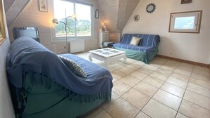 Une pièce mansardée chaleureuse, avec un canapé bleu, une table basse blanche et une vue sur l’extérieur grâce à une grande fenêtre.