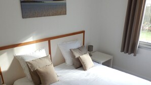 Ein ordentlich bezogenes Bett mit weißer und beiger Bettwäsche, ein hölzerner Kopfteil und ein Nachttisch mit einer Lampe. Über dem Bett hängt ein Bild von einer Strandlandschaft.