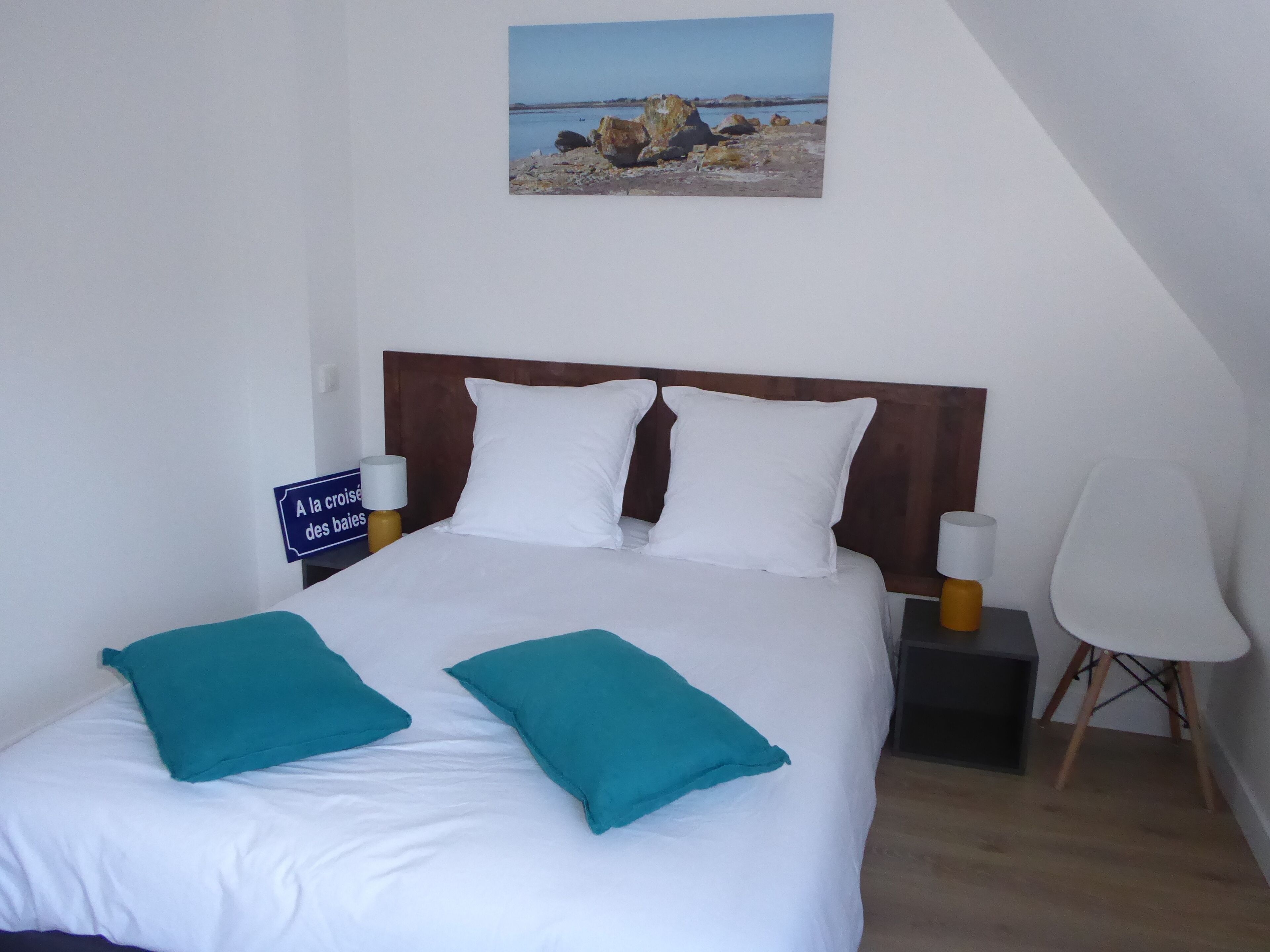 A neatly made bed with white and teal pillows, a wooden headboard, and a nightstand with a lamp. A painting of a rocky beach is on the wall above the bed.