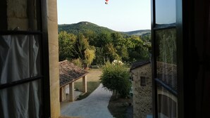 Vista pela janela de um edifício com fachada de pedra, área externa coberta e um caminho que leva a uma colina arborizada.