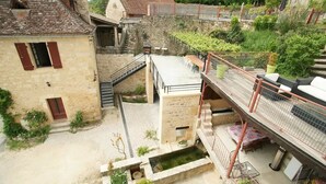 Um pátio com uma escadaria de pedra que leva a uma área coberta, cercado por edifícios de pedra com persianas vermelhas.