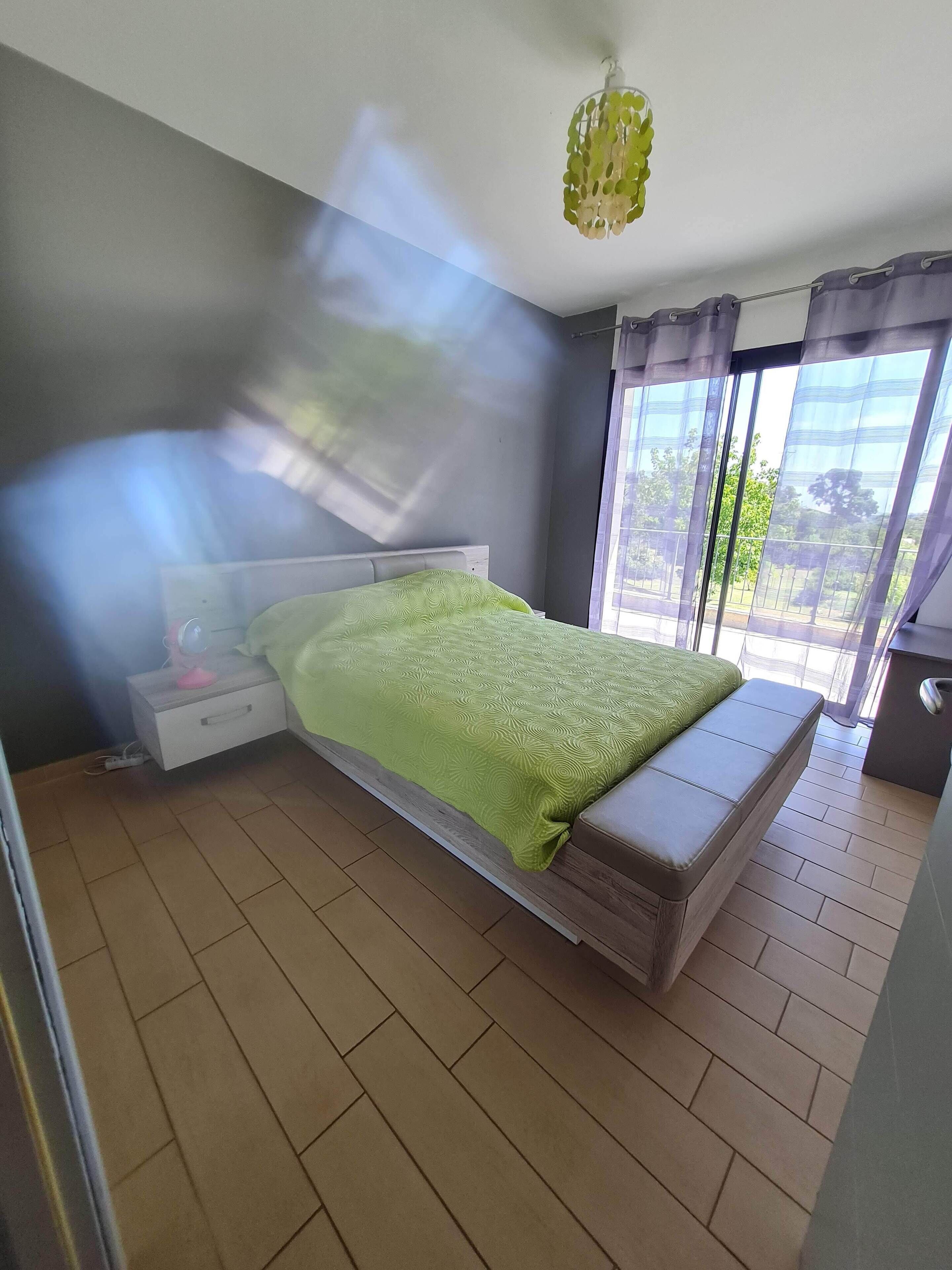 A bedroom with a bed, a bedside table, a nightstand, a window with curtains, and a view of the outdoors.