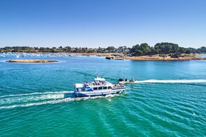 Un barco con pasajeros en un mar tranquilo, amarrado cerca de una costa con edificios y árboles.