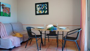 A dining area with a table set for two, a sofa with a hat on it, and a framed abstract painting on the wall. - Apartment in the heart of Vaucluse close to the tourist attractions of Vaucluse (Loriol du Comtat)