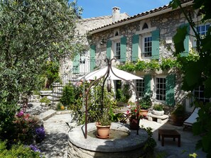 A courtyard with a fountain, potted plants, and a stone building with green shutters. - Vacation rental villa pool Orgon LS1-117 (Orgon)