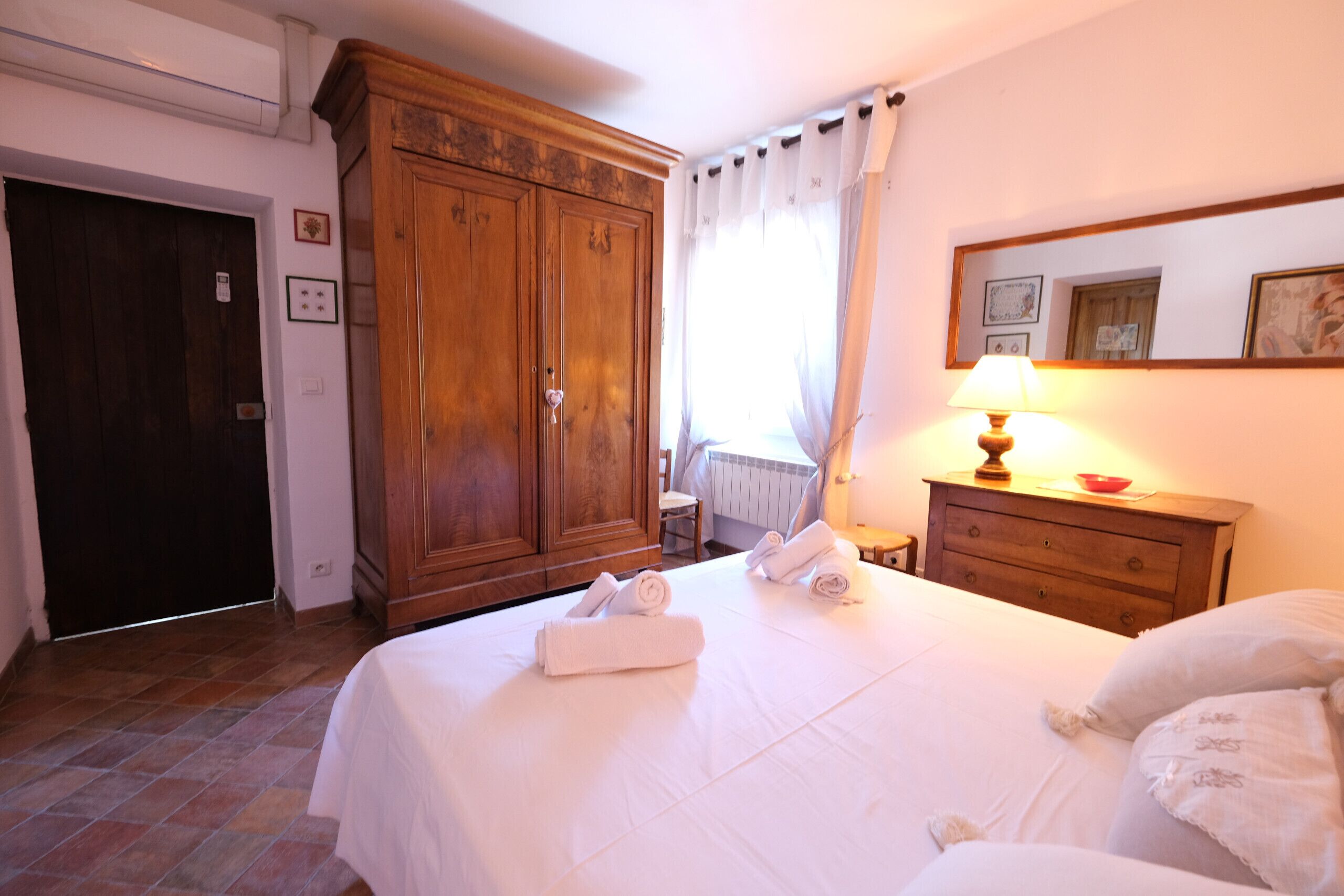A hotel room with a large wooden wardrobe, a bed with white linens, a bedside table with a lamp, and a window with curtains.