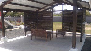 A covered patio area with two wooden chairs and a table, a stone floor, and a view of a garden and stairs in the background.