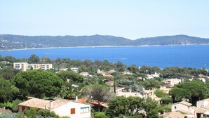 Un pueblo costero con edificios rodeados de árboles, un mar azul cristalino y colinas al lejano horizonte.