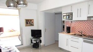 Cocina compacta con armarios blancos, microondas y cafetera. Hay un televisor sobre un pedestal de vidrio y una mesa de comedor con sillas.