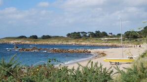 A beach with a rocky shoreline, a yellow boat, and a grassy hill with trees in the background. - Kervegan magnifique villa vue mer à Pleumeur Bodou (PLEUMEUR BODOU)