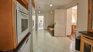 Une cuisine avec des meubles de rangement en bois, un four blanc et un plan de travail noir. On y trouve une poubelle et une porte qui donne sur une autre pièce.