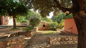 Un sentier de pierres mène à une construction rustique en pierre, dotée d’une petite ouverture, et est entouré d’arbres et d’un mur de pierres.