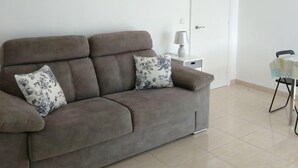 Una sala de estar con un sofá marrón, dos cojines estampados, una mesita de noche con una lámpara y un ventilador de techo.