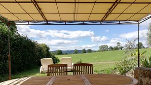 Un espacio de estar techado al aire libre con una mesa y sillas de madera, con vista a un campo verde y colinas distantes.