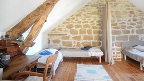 Un dormitorio acogedor en el ático, con techo inclinado, pared de piedra, vigas de madera, una cama, una mesita pequeña y una silla.
