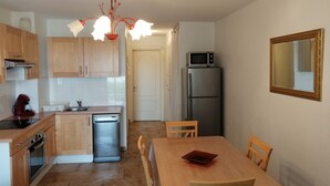 Cocina compacta con armarios de madera, lavavajillas, microondas y refrigerador. Hay una mesa de comedor con sillas y un solo libro sobre ella.