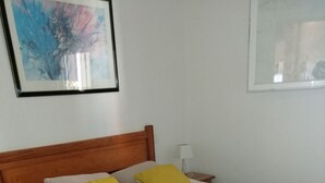 Una cama con cubrecama floral, dos almohadas, cabecera de madera, mesita de noche con lámpara y un cuadro abstracto enmarcado en la pared.
