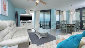 Amplia sala de estar con un sofá blanco, una mesa de centro y un sillón colorido. Hay un televisor de pantalla plana, una zona de comedor con sillas y un balcón con vistas.