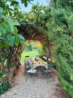 An outdoor dining area with a table set for four, surrounded by greenery and a pergola overhead.