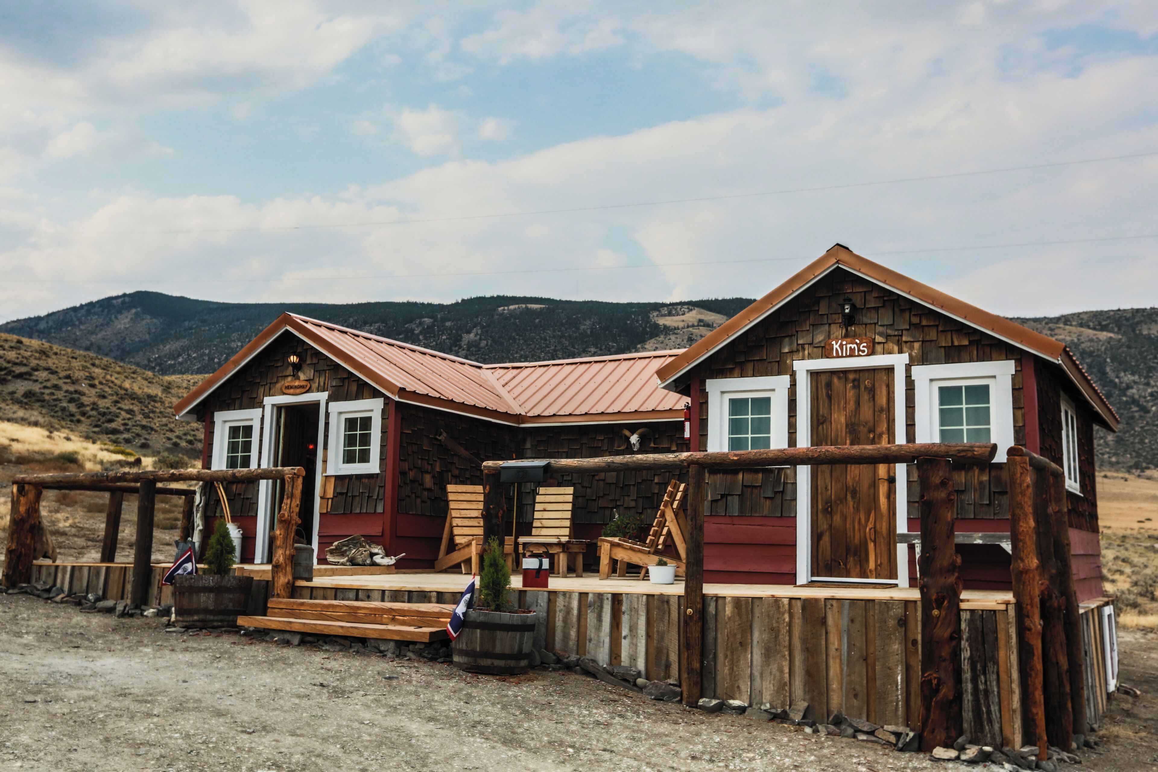 Une maison rustique en bois, à l’extérieur rouge et au toit métallique, dotée d’un porche avec des chaises en bois et une petite table.