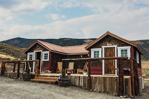 Een rustiek houten huis met een rode gevel en een metalen dak, met een veranda met houten stoelen en een kleine tafel.