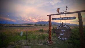 Ein rustikales Holzschild mit der Aufschrift "Walking Star Ranch" und "Apple Jack Ranch" vor einem Sonnenuntergangshimmel und fernen Bergen.
