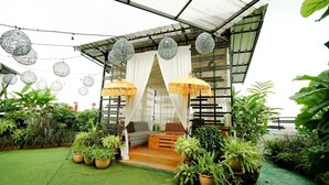 A covered outdoor seating area with a wooden bench, white curtains, and hanging spherical decorations. - Deli Hotel (Medan)