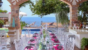 An outdoor dining area with a view of the sea, featuring a glass table set with pink and white tableware, surrounded by white chairs.